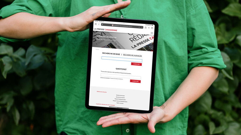 Une personne tient une tablette devant sa poitrine. L’écran affiche la page de recherche du Guide de rédaction en ligne de La Presse Canadienne.