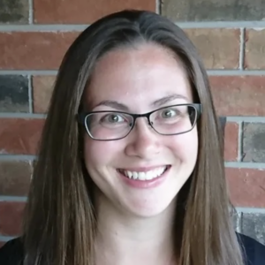 Portrait d'Amanda Feeney. Amanda a les cheveux longs bruns et le teint clair. Elle porte des lunettes à monture foncée. Elle est debout devant un mur de briques.