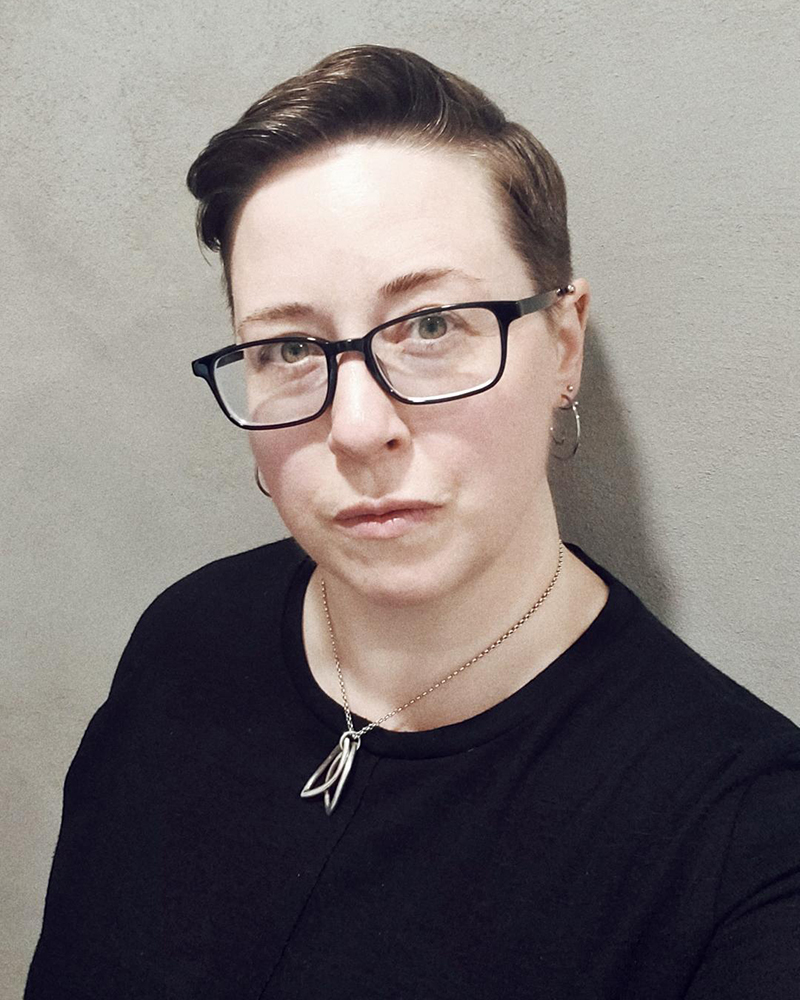 Portrait de Andrea Zanin. Andrea est une personne blanche avec les cheveux bruns courts qui porte des lunettes à monture noire, un t-shirt noir et des bijoux en argent.